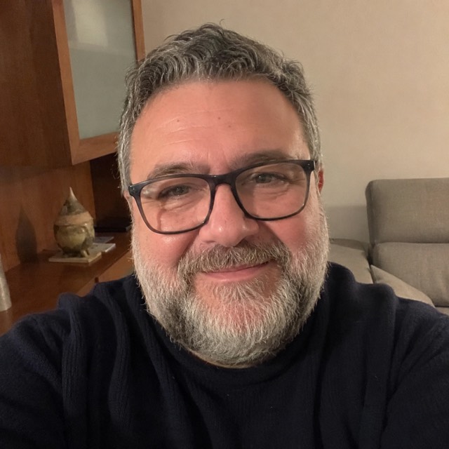 Ritratto di un uomo italiano robusto sulla sessantina, capelli grigi, occhiali scuri e barba grigia, maglione scuro, in salotto