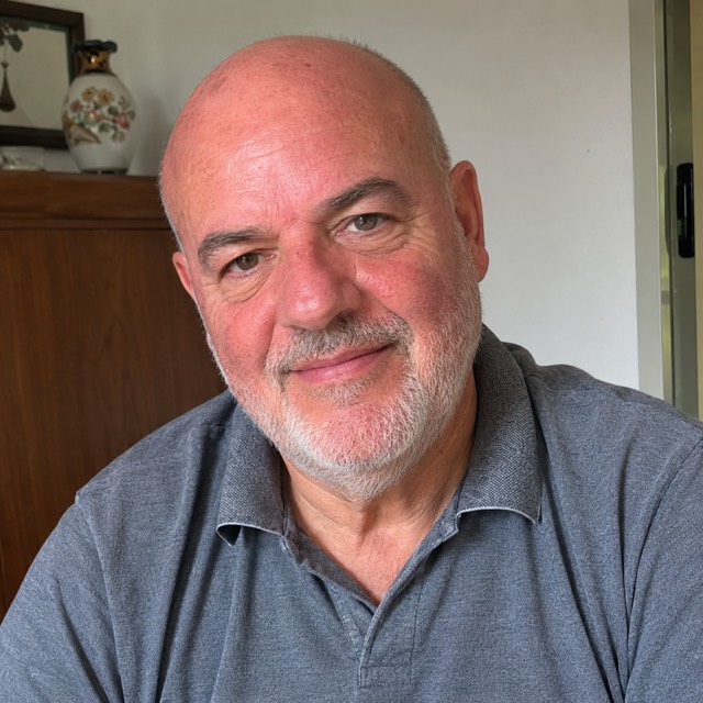 Ritratto di un uomo italiano sui sessant'anni, calvo con barba brizzolata corta, polo grigia, in casa