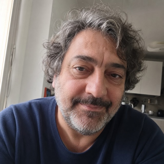 Ritratto di un uomo italiano sulla sessantina, capelli grigi ricci e barba grigia, carnagione olivastra, maglione blu, in casa